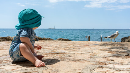 A baby with sunscreen clothes looks at the sea on vacation