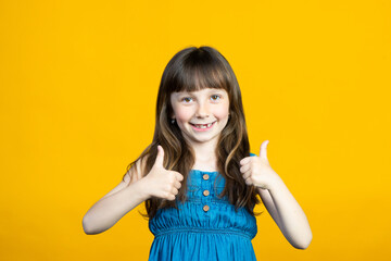 A little smiling girl raised her finger thumb up and shows Cool, Super. Lost milk teeth in the mouth. Dentist concept. Isolated on yellow background