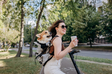 Happy smiling woman traveler is standing with electro scooter and drinking coffee in city parkland with dog Welsh Corgi Pembroke in a special backpack