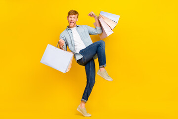 Full size photo of handsome lucky guy dressed blue shirt denim jeans hold bags celebrating new clothes isolated on yellow color background