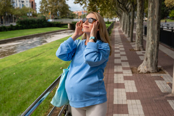 Pregnant woman looks at the sun in sunglasses. Mom-to-be in a blue hoodie and beige leggings walks around the city