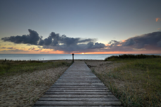 Ein Holzweg f&uuml;hrt zum Strand von Braderup auf Sylt, wo gerade die Sonne aufgeht und ein kleiner Regenschauer f&auml;llt.