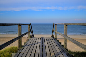 Fototapeta na wymiar Von einer Holzbrücke hat mein ein Panorama Blick auf das Meer und den Sandstrand. Eine Treppe führt hinab zum Strand.