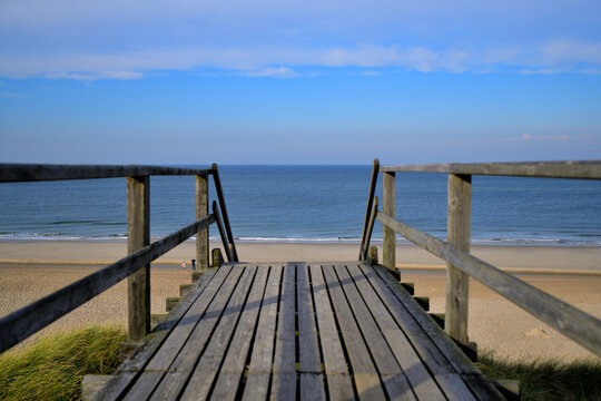 Von einer Holzbr&uuml;cke hat mein ein Panorama Blick auf das Meer und den Sandstrand. Eine Treppe f&uuml;hrt hinab zum Strand.