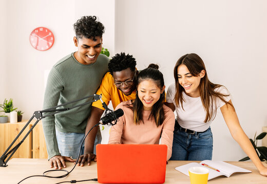 Happy group of multiracial young student friends working together recording radio podcast using microphone and laptop at home office. Teamwork and community concept with diverse people live streaming