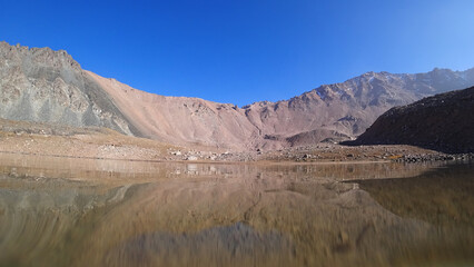 Transparent mountain lake surrounded by rocks. The water is like a mirror, reflecting the rocks and the slope. Light ripples on the surface of the water. The bottom is visible. There are big stones