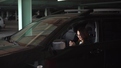 A young businesswoman uses a phone while sitting in a car in a parking lot. A woman in glasses and a jacket is texting on a smartphone.