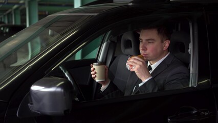 Fototapeta na wymiar A young businessman is having lunch in a car in a parking lot. A man in a suit eats fast food and enjoys the taste. The businessman eats a cheeseburger and drinks coffee.