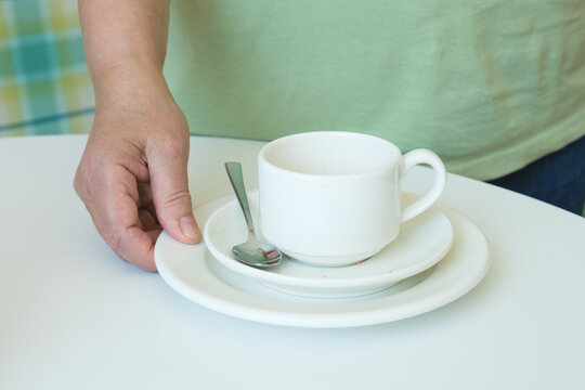 Close-up Of A Hand Picking Up A Stack Of Plates And A Dirty Coffee Cup From The Table. The Waiter In The Cafe Cleans The Dirty Dishes From The Table. Cafeteria Maintenance Work.