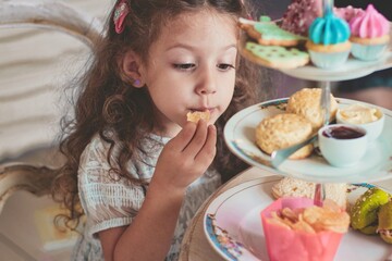 A cute little girl enjoying high tea with ceramic tiered plate stand with scones, cupcakes, sandwiches and crisps - obrazy, fototapety, plakaty