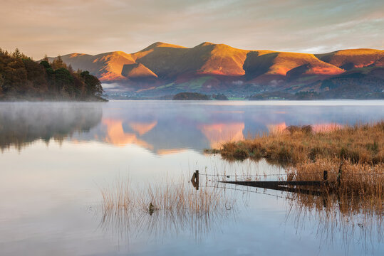 Absolutely stunning vibrant Autumn sunrise landscape image looking from Manesty Park in Lake Distict towards sunlit Skiddaw Range with mit rolling across Derwentwater surface