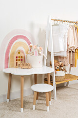 Children's room with Montessori Clothing Rack, white table and Storage Baskets with toys. Dress, jacket and sweaters on hangers in wardrobe. Nursery Storage Ideas. Montessori Toddler Room.