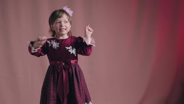 Cute little girl in an vintage burgundy dress is dancing, rejoicing and making funny faces. The concept of children theater productions and developing acting circles. Pink background