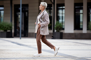 Side view full length woman walks outside and wears trendy business clothes, oversize jacket, tie, pants and white loafer shoes. Confident female model with short blonde hair