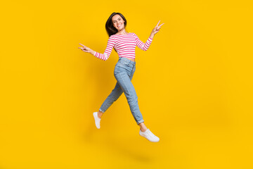 Fototapeta na wymiar Full size photo of gorgeous good mood girl wear striped long sleeve jeans showing v-sign jump fly isolated on yellow color background