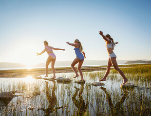 Girls (10-11, 12-13, 14-15) walking on stepping stones in lake at sunrise