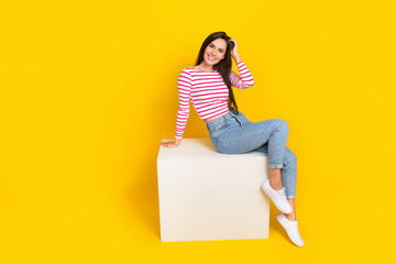 Photo of sweet lovely nice girl long hairdo wear striped shirt jeans sitting on cube arm touch hair isolated on yellow color background