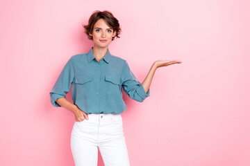 Portrait photo of young confident pretty smile woman put hand pocket hold palm showing new product recommend information isolated on pastel pink color background