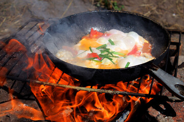 fried eggs on a campfire in the forest, fried eggs on a campfire on a camping trip, scrambled eggs in a frying pan on fire, cook an omelet on a fire, Cooking on a campfire, breakfast on the fire - obrazy, fototapety, plakaty
