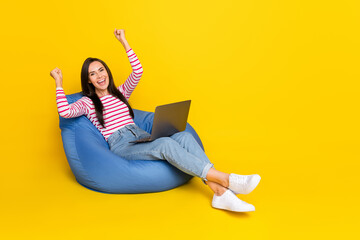 Full size photo of cheerful girl striped shirt jeans sit in pouf with laptop scream yeah fist up...