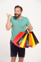 Bearded smiling man holding shopping bags and pointing by thumb up to copy space over white background and looking at camera