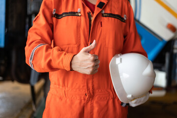 A repairman in orange coverall is thumbing up and holding white safety helmet with blurred background of garage workshop. Safe working practice in industrial concept.