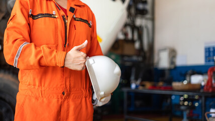 A repairman in orange coverall is thumbing up and holding white safety helmet with blurred...