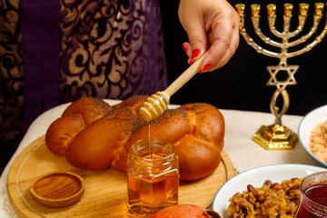 Obraz na płótnie Canvas Honey drips from a wooden spoon in a woman's hand next to the menorah challah and couscous and pomegranate.