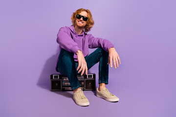 Full body photo of cool beard red hairdo guy sit listen boombox wear sunglass hoodie jeans shoes isolated on purple background - obrazy, fototapety, plakaty