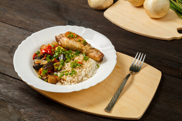 Rice with vegetables and chicken leg on a handmade wooden board next to a fork on a wooden table.