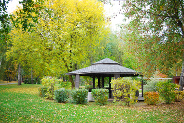 Landscape in the autumn park. Wooden gazebo for relaxing among yellow foliage and autumn trees. Wooden gazebo in the autumn forest among fallen leaves.