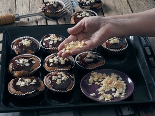 The hand of an elderly woman sprinkles freshly baked homemade muffins, poured with chocolate icing, coconut flakes.