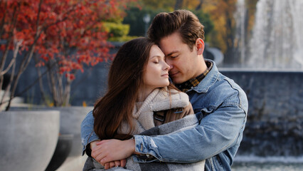 Fototapeta premium Caucasian couple lovers partners stand background of city fountain pressed closely to cheeks together spend time in autumn park on date embrace feel love emotion romantic atmosphere happy young family