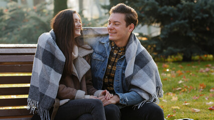 Cute loving couple sit on bench warming each other covering wool blanket outdoors in cold froze autumnal weather feel care love hugging funny talking with steam from mouth enjoy nature romantic date