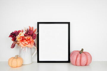 Mock up black picture frame with pink hue autumn flowers and pumpkins on a white shelf against a...