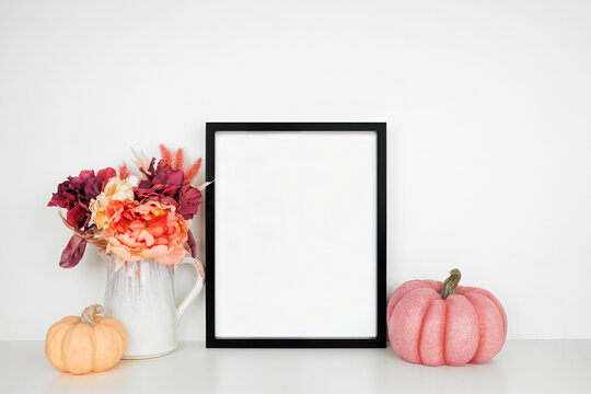 Mock up black picture frame with pink hue autumn flowers and pumpkins on a white shelf against a white wall. Fall concept. Copy space.