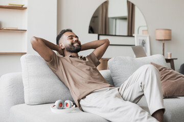 Satisfied handsome young man relaxing on sofa at home in living room, resting after a hard day work, closed eyes, put hands behind head, smiles happily. Relaxation, self care, enjoy life concept - obrazy, fototapety, plakaty