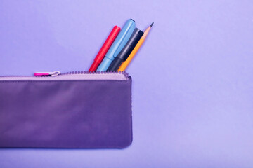 Fashionable violet pencil case with multicolored felt-tip pens, pencils and pens on violet background.
