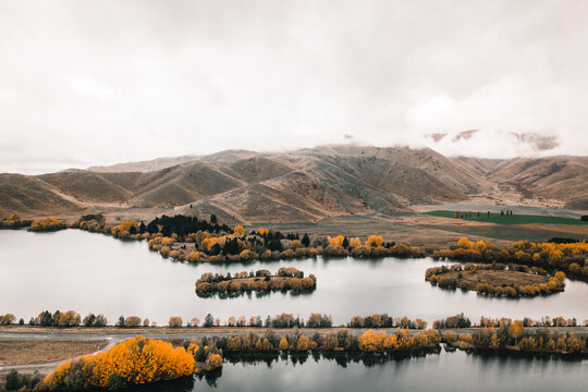 Lonely And Narrow Straight Road That Crosses Between Two Lakes In The Middle Of Vegetation And Trees Of Different Colors Next To Many Mountains On A Cloudy And Stormy Day, Mount Cook, New Zealand