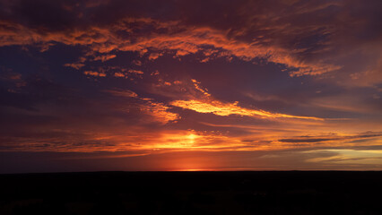 Clouds reflecting sunlight after sun has set below horizon, in hues of pink and orange over silhouetted, dark earth