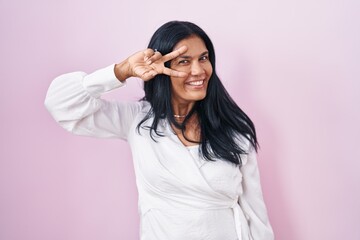 Mature hispanic woman standing over pink background doing peace symbol with fingers over face,...