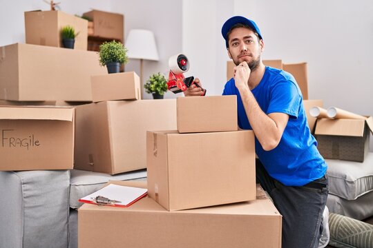 Hispanic man with beard working moving boxes serious face thinking about question with hand on chin, thoughtful about confusing idea