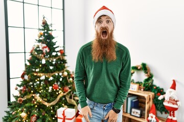 Redhead man with long beard wearing christmas hat by christmas tree afraid and shocked with surprise and amazed expression, fear and excited face.