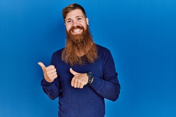Redhead man with long beard wearing casual blue sweater over blue background pointing to the back...