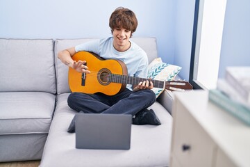 Young blond man having online classical guitar class sitting on sofa at home