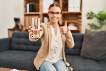 Young caucasian woman offering a glass of water smiling happy and positive, thumb up doing excellent and approval sign