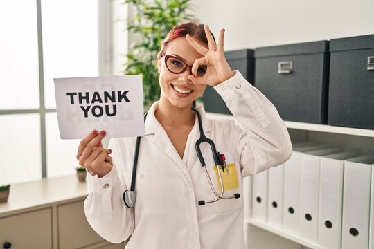 Young caucasian woman wearing doctor uniform holding thank you banner smiling happy doing ok sign with hand on eye looking through fingers