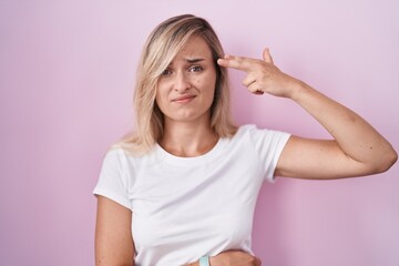 Young blonde woman standing over pink background shooting and killing oneself pointing hand and fingers to head like gun, suicide gesture.