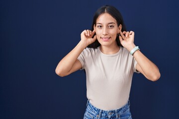 Young hispanic woman standing over blue background smiling pulling ears with fingers, funny...