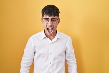 Young hispanic man standing over yellow background angry and mad screaming frustrated and furious,...